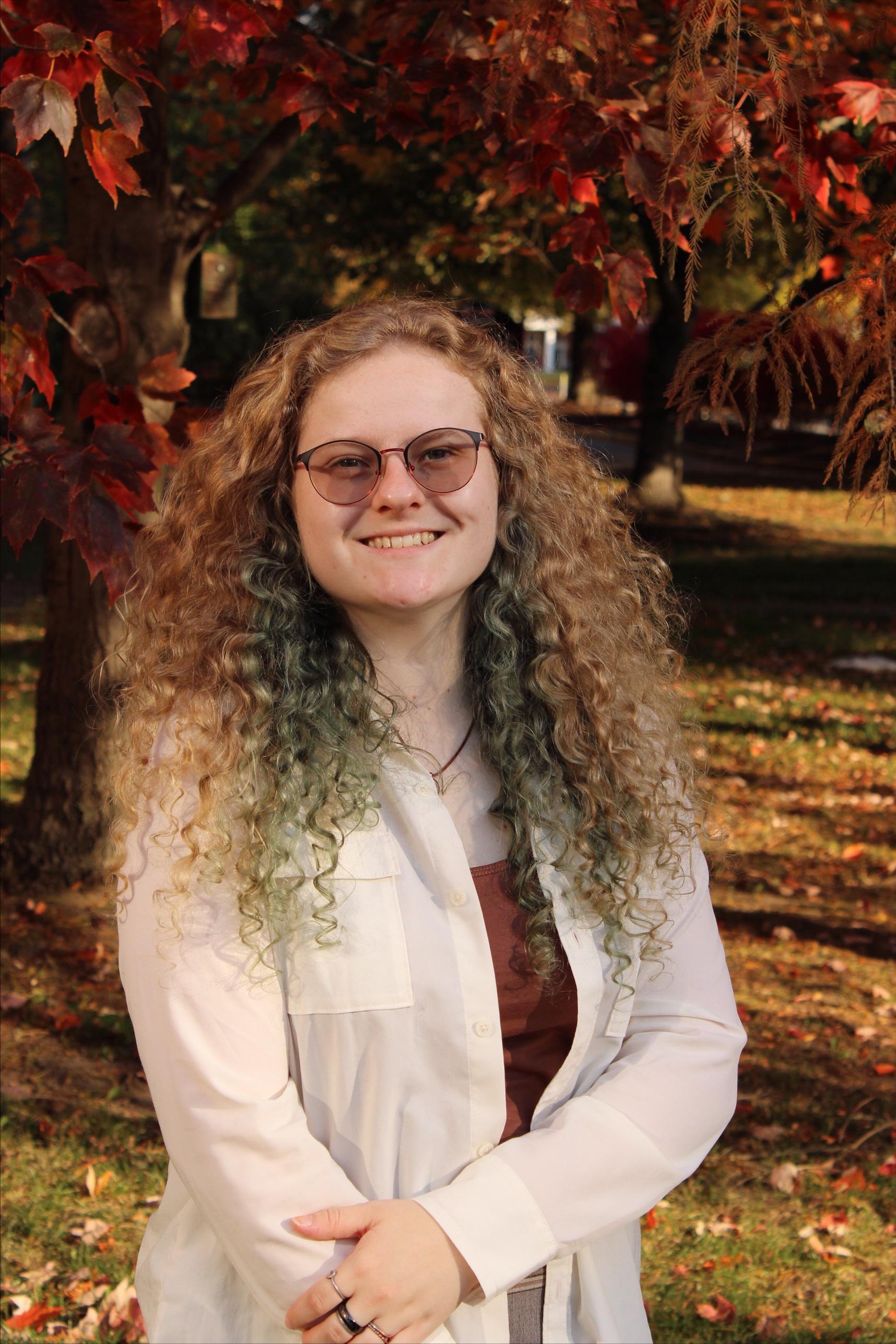 Arianne, smiling and wearing a white shirt with fall foliage behind her. 